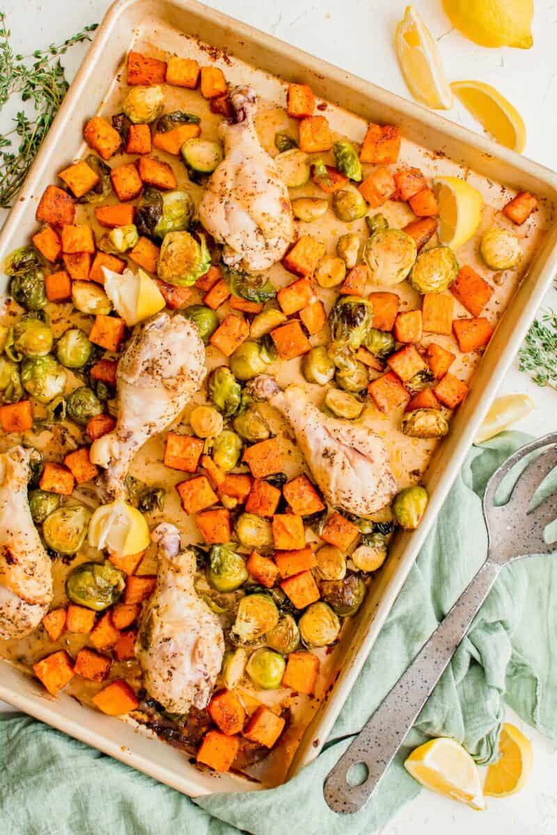 lemon pepper drumsticks and roasted vegetables on a rimmed metal sheet pan next to fresh lemon slices