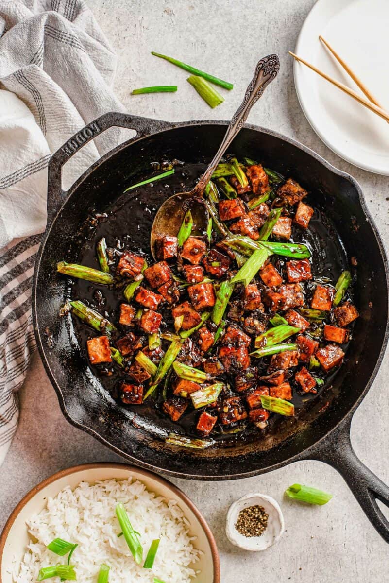 Tofu cubes are presented in a black pan with green onions. 