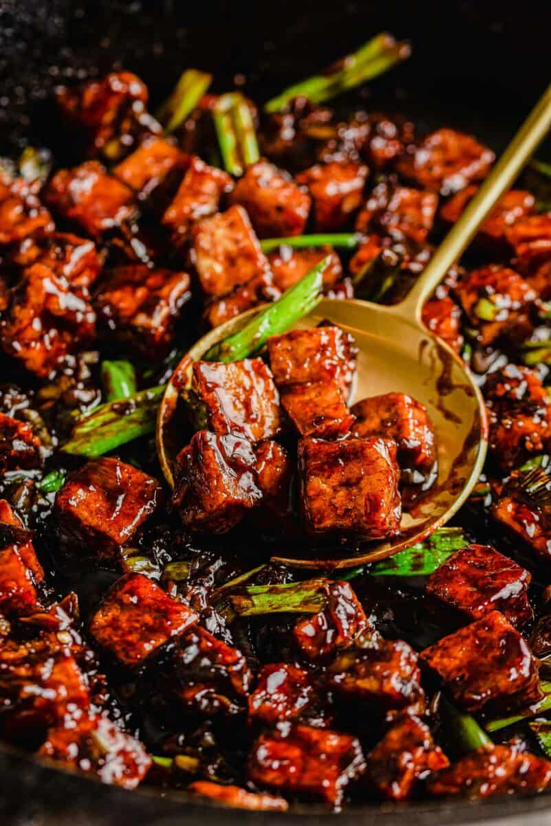 A serving spoon is scooping up a portion of cooked tofu. 