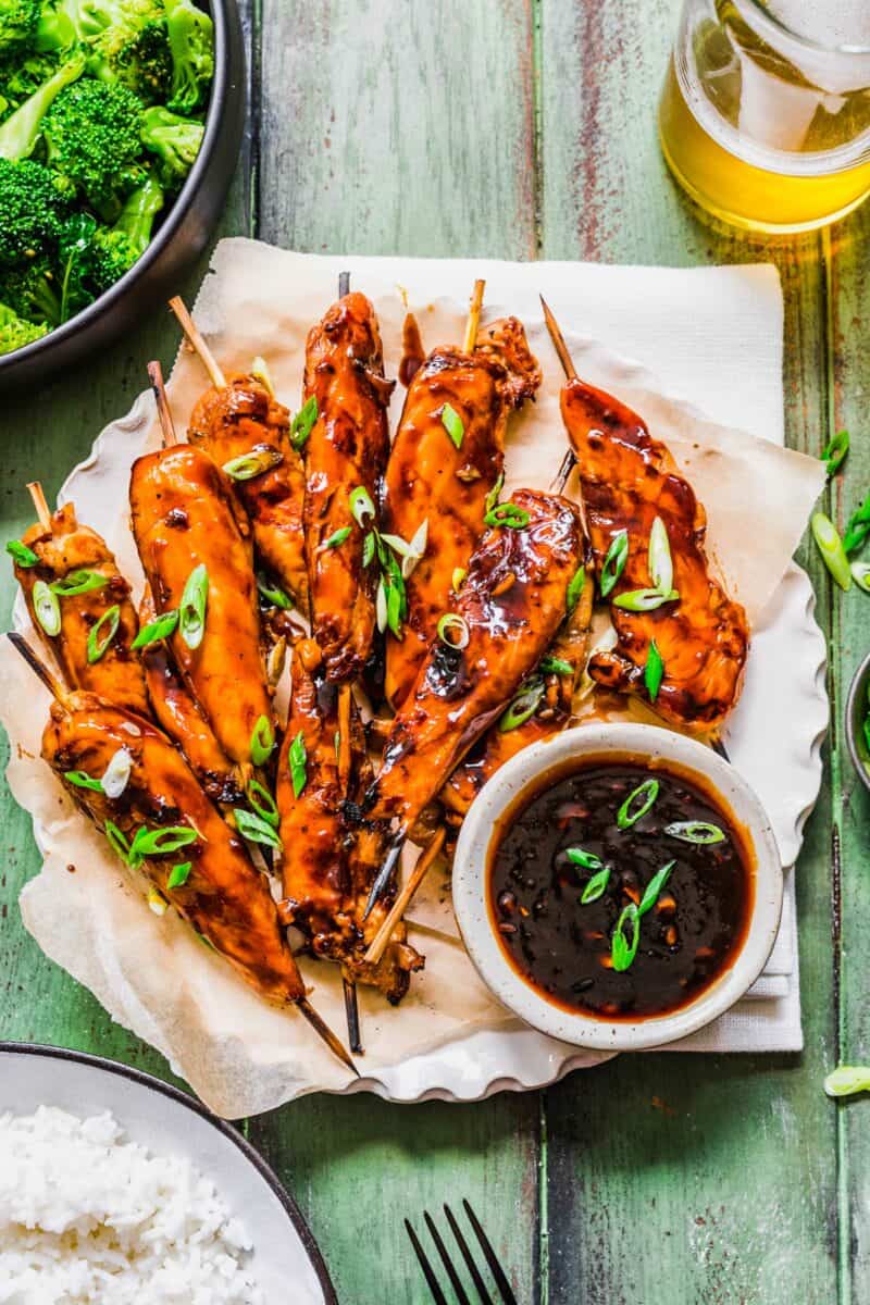 A bowl of teriyaki sauce is plated next to a large serving of grilled chicken skewers. 