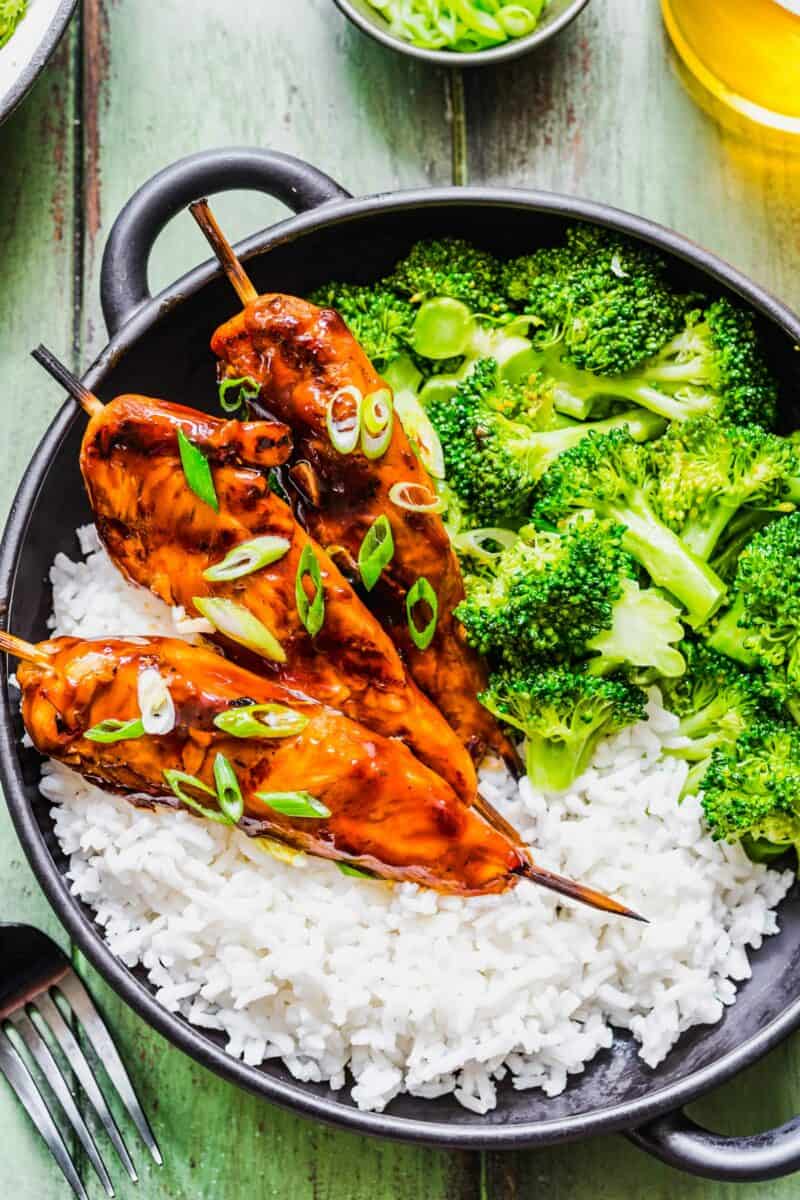 White rice is placed in a bowl with steamed broccoli and chicken skewers. 