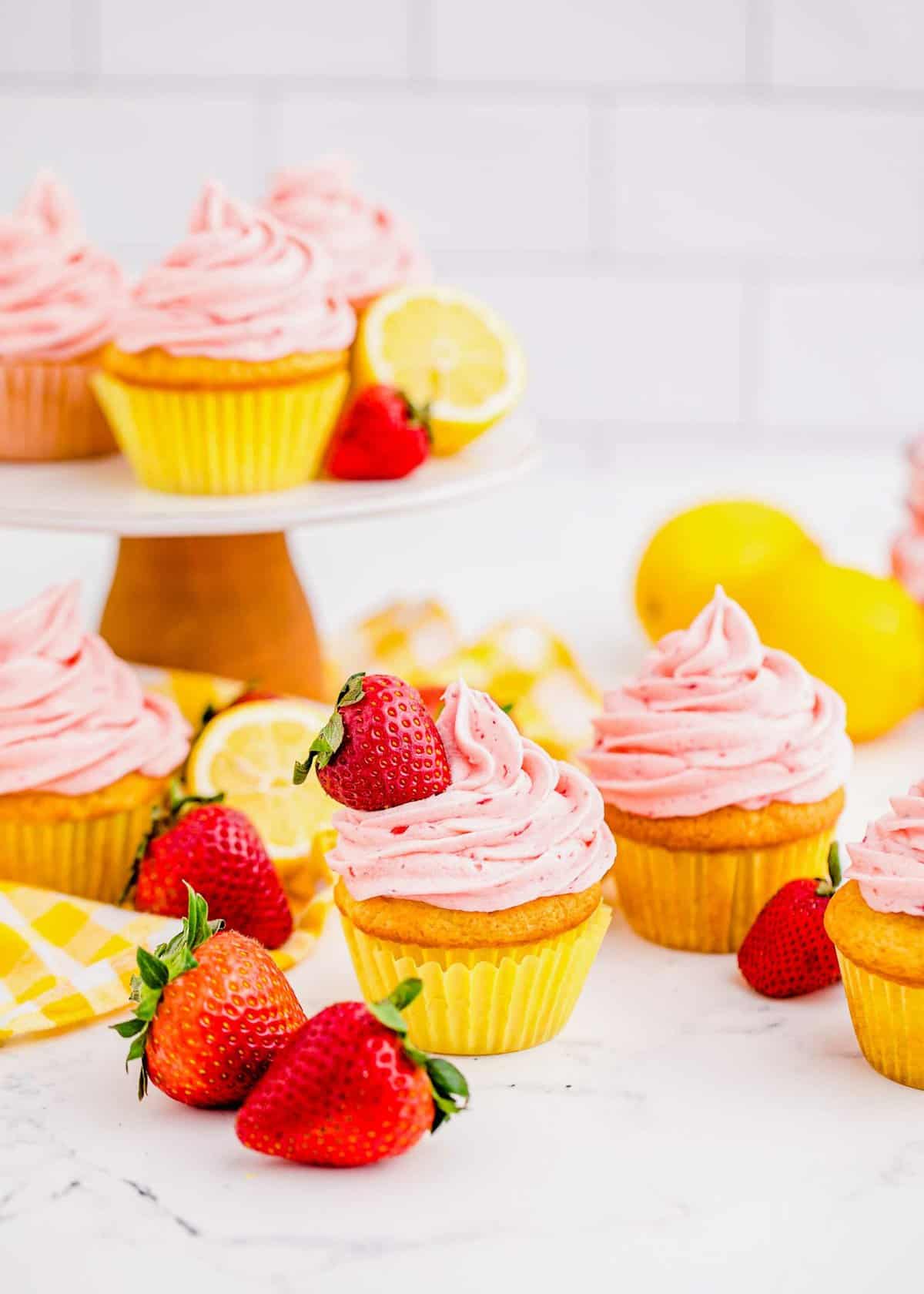 Strawberry lemonade cupcakes on countertop and cake stand, with fresh strawberries