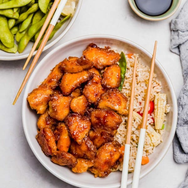 overhead image of sweet and sour chicken in a bowl on top of vegetable fried rice with wooden chopsticks resting on top. a plate of edamame and wooden chopsticks can be seen next to the sweet and sour chicken