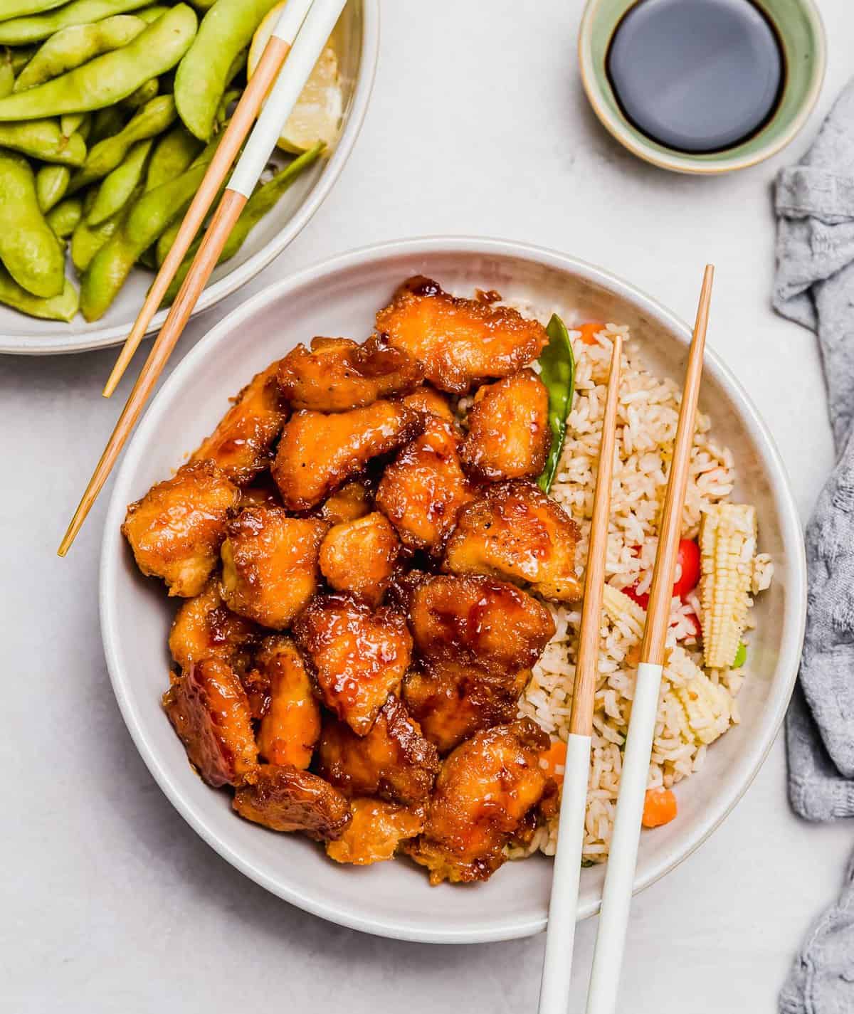 overhead image of sweet and sour chicken in a bowl on top of vegetable fried rice with wooden chopsticks resting on top. a plate of edamame and wooden chopsticks can be seen next to the sweet and sour chicken