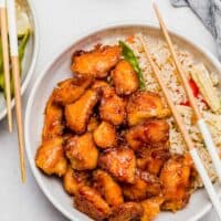 overhead image of sweet and sour chicken in a bowl with fried rice and wooden chopsticks resting on the bowl
