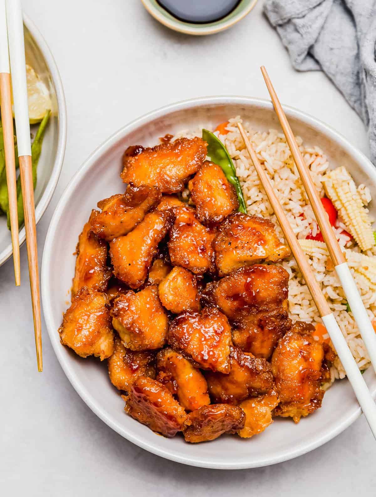 overhead image of sweet and sour chicken in a bowl with fried rice and wooden chopsticks resting on the bowl