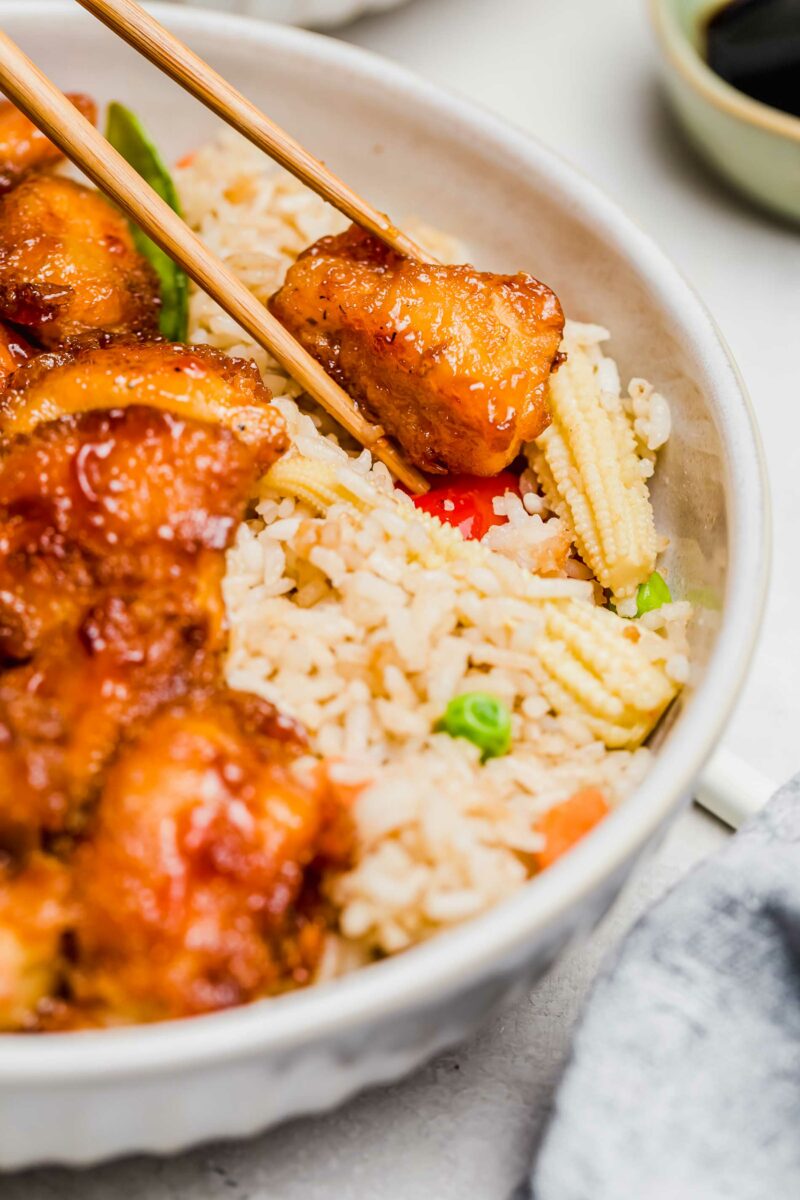 up close image of sweet and sour chicken on top of fried rice with wooden chopsticks picking up the piece of chicken