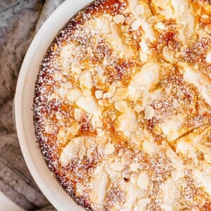 A golden brown Apple Almond Coffee Cake sits on a white plate. It is topped with almond slices and finished with powdered sugar.