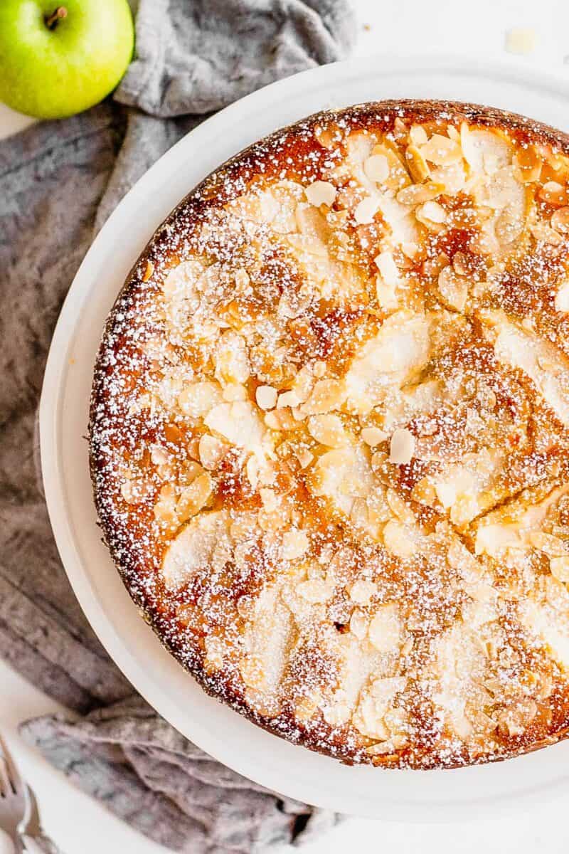 A whole Apple Almond Cake with lightly toasted almonds and sprinkled with powdered sugar sits on a white plate. A gray cloth napkin and whole apple sit next to the cake.