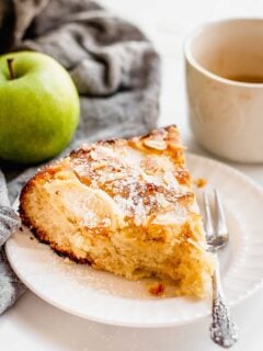 One slice of Almond Apple Cake sits on a white plate with a fork next to it. A whole Granny Smith apple and a cup of coffee sits next to the cake slice.