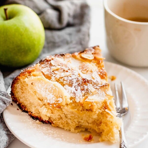 One slice of Almond Apple Cake sits on a white plate with a fork next to it. A whole Granny Smith apple and a cup of coffee sits next to the cake slice.