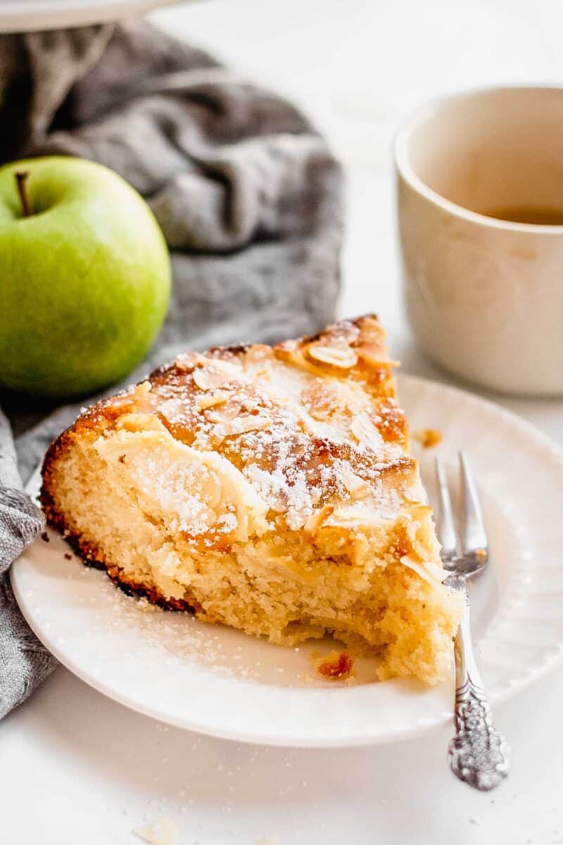 Slice of Apple Almond Cake served on a white dessert plate sits next to a fork. A cup of coffee and a whole apple is in background.