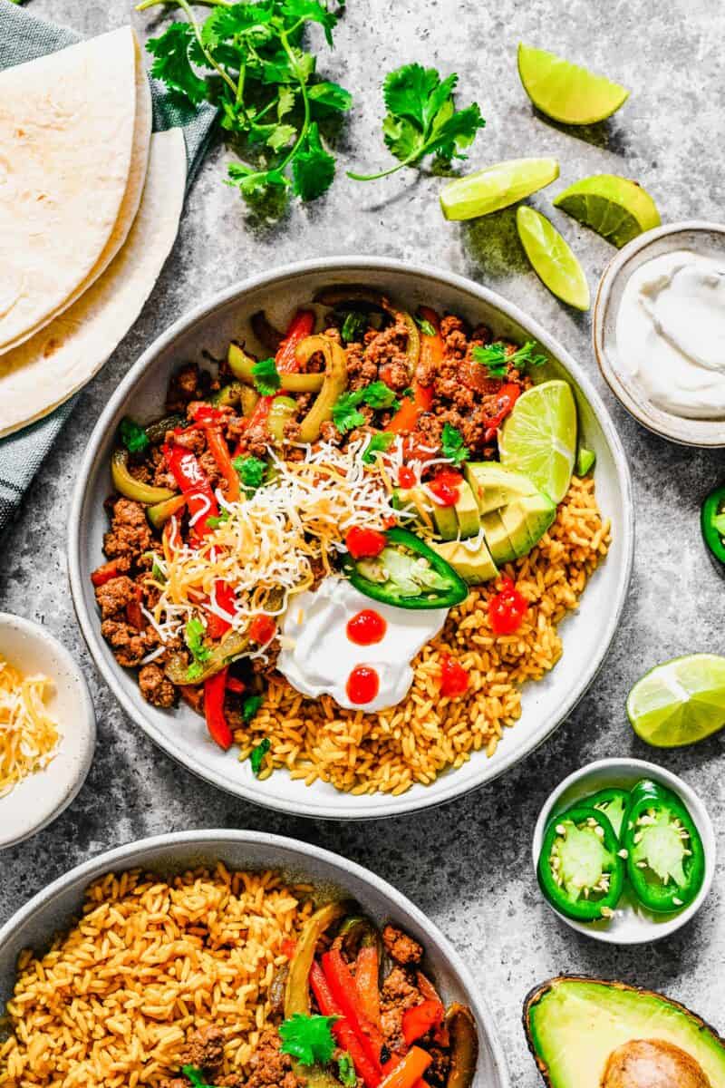 A white bowl is filled with ground beef fajitas, rice, and several toppings. 