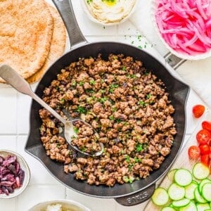 A skillet of Middle Eastern inspired ground beef is surrounded by pita bread, hummus, pickled onions, cherry tomatoes, cucumber slices, olives, and a bowl of rice on a white surface.