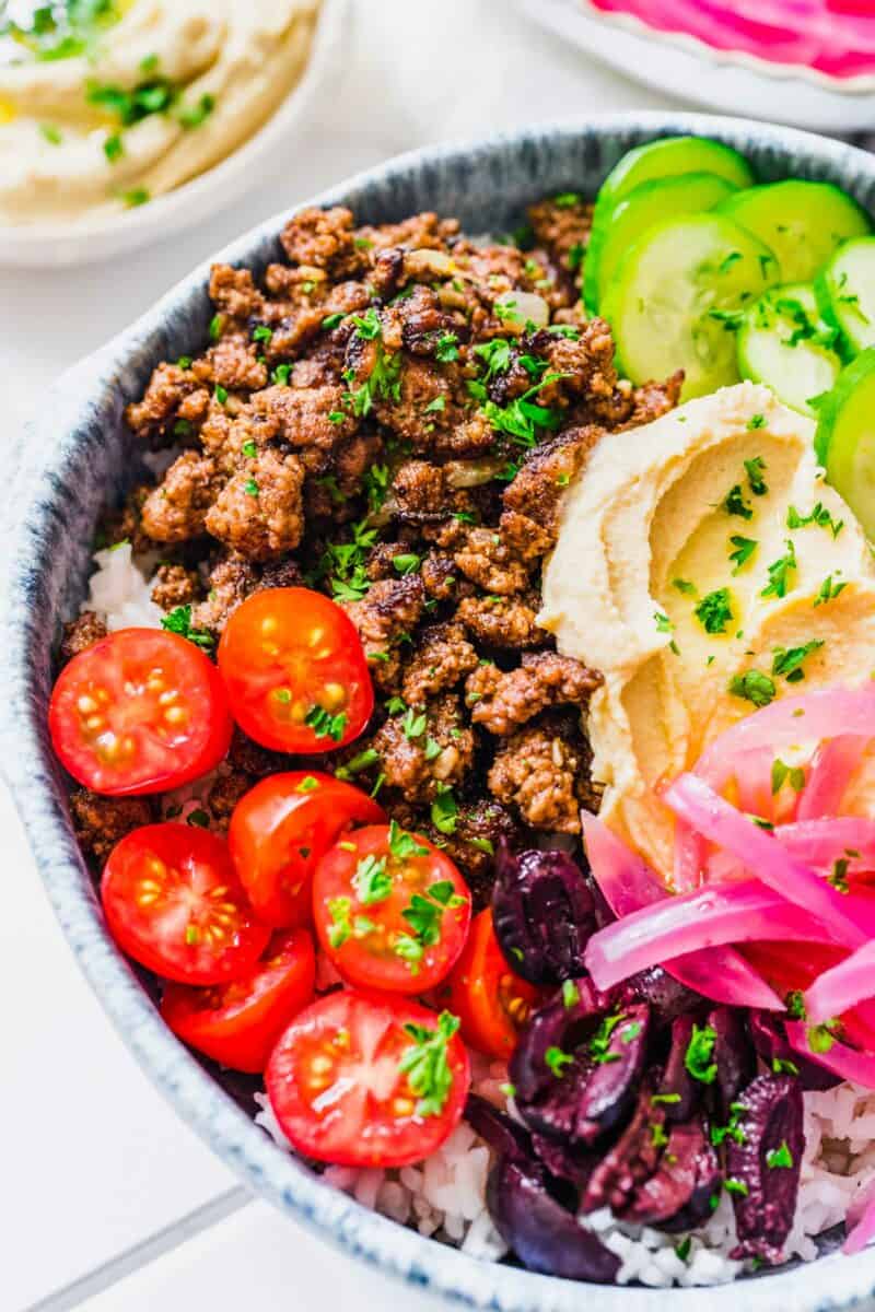 Tomatoes, olives, and pickled red onions are all placed on top of ground beef and rice in a bowl. 