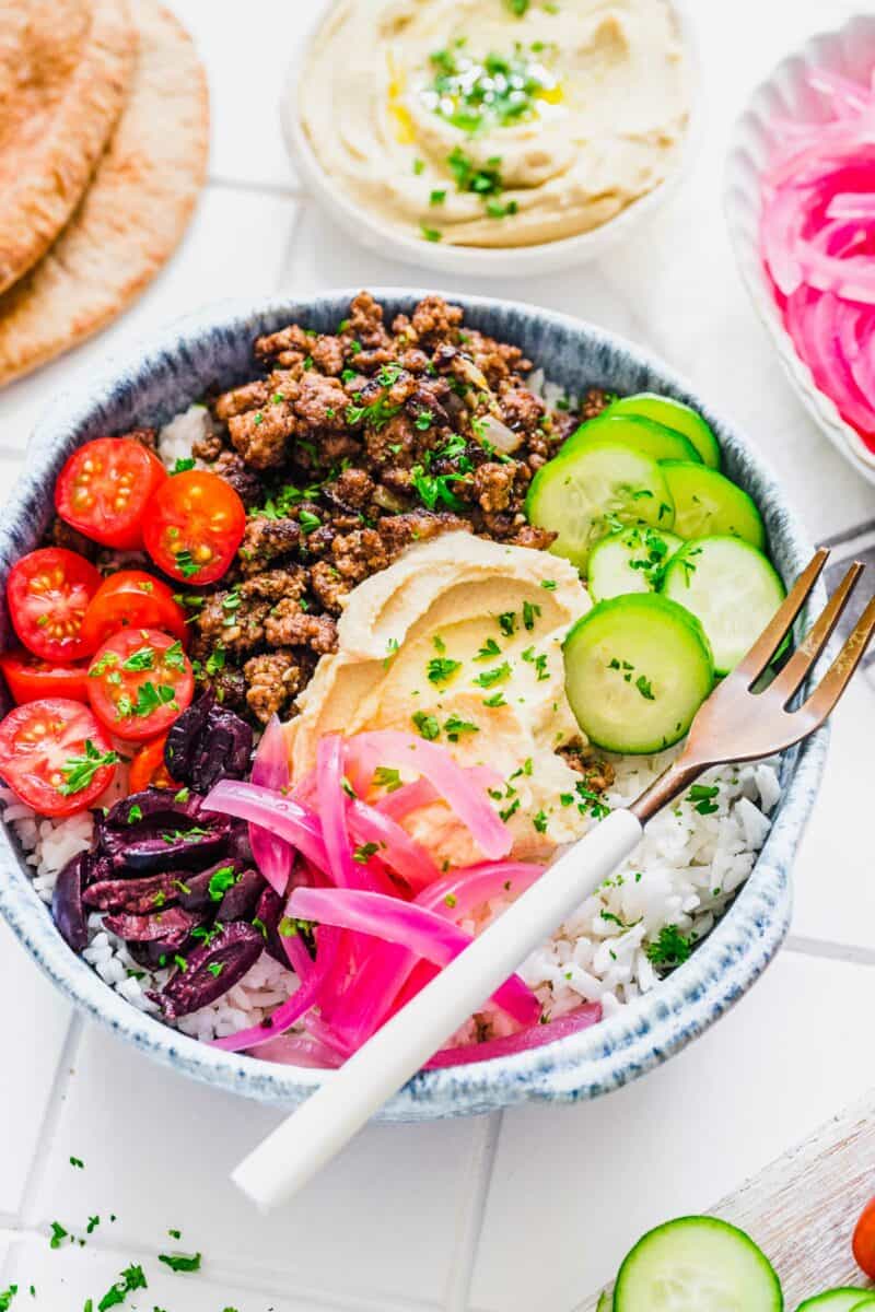 Fresh veggies are placed on top of a full beef bowl with rice and hummus.