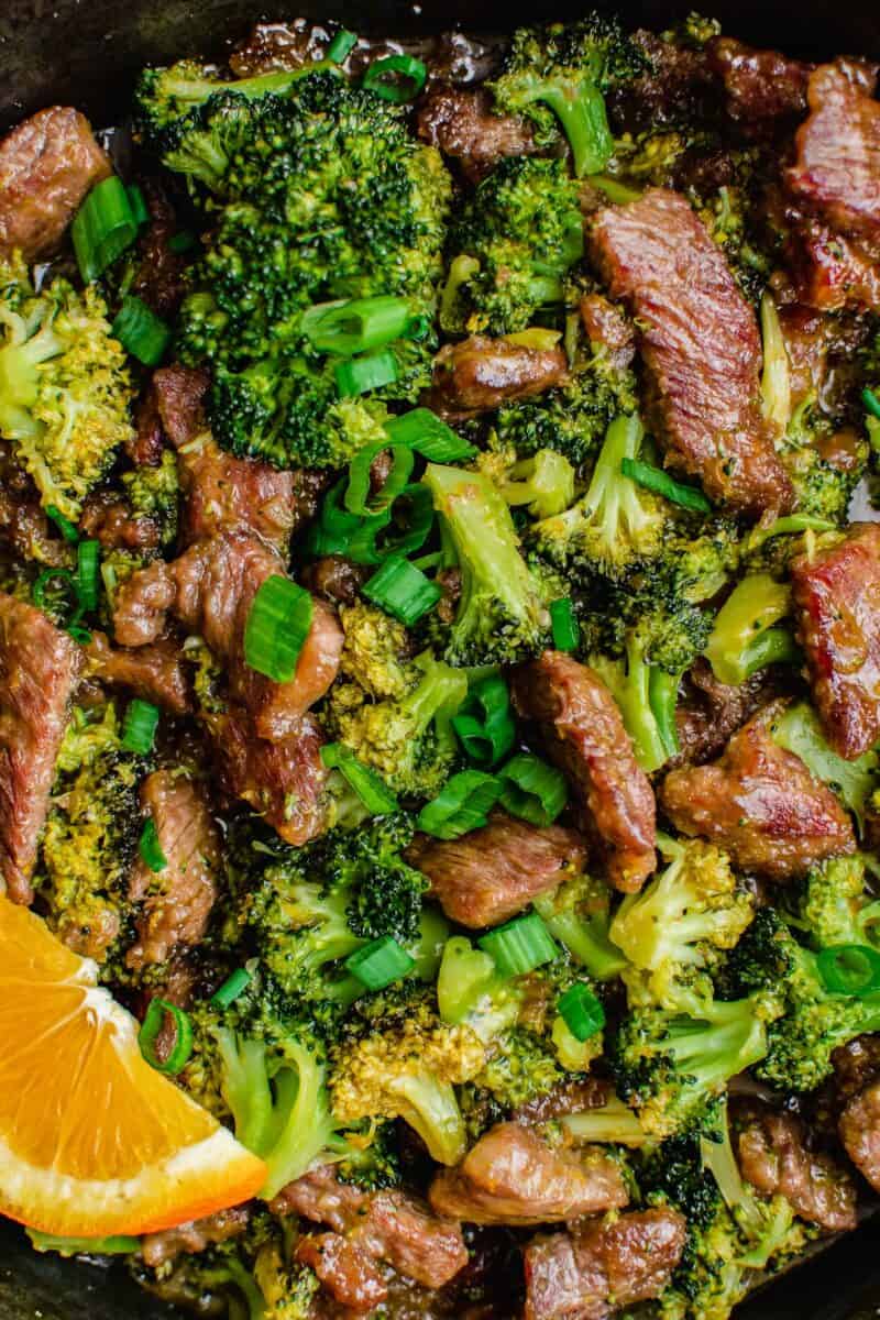 Cooked beef and broccoli is garnished with green onions and a single orange slice. 