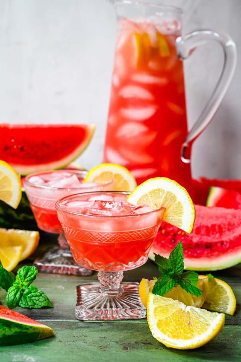 Two glasses of watermelon lemonade are garnished with lemon wedges. 