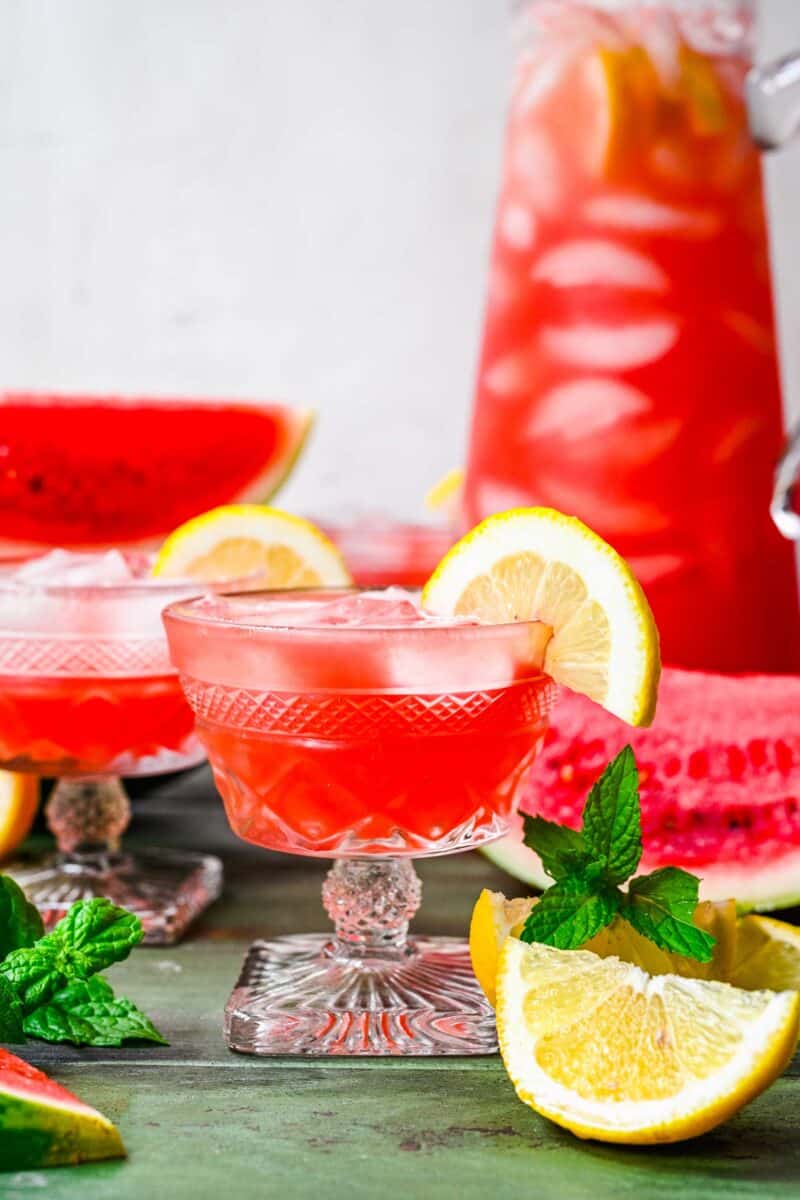 Lemon wedges and mint sprigs are placed around several glasses of watermelon lemonade. 