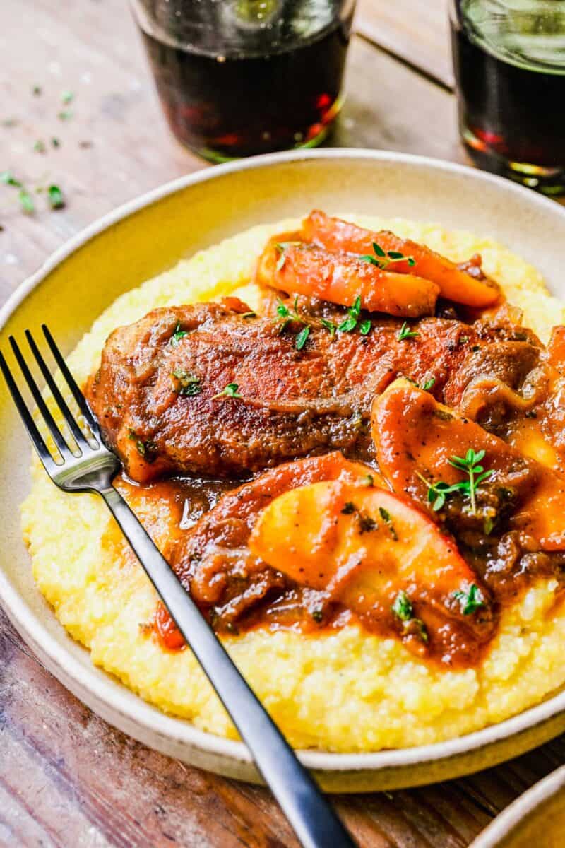 A fork is placed on a round white plate filled with apple butter pork chops and polenta. 