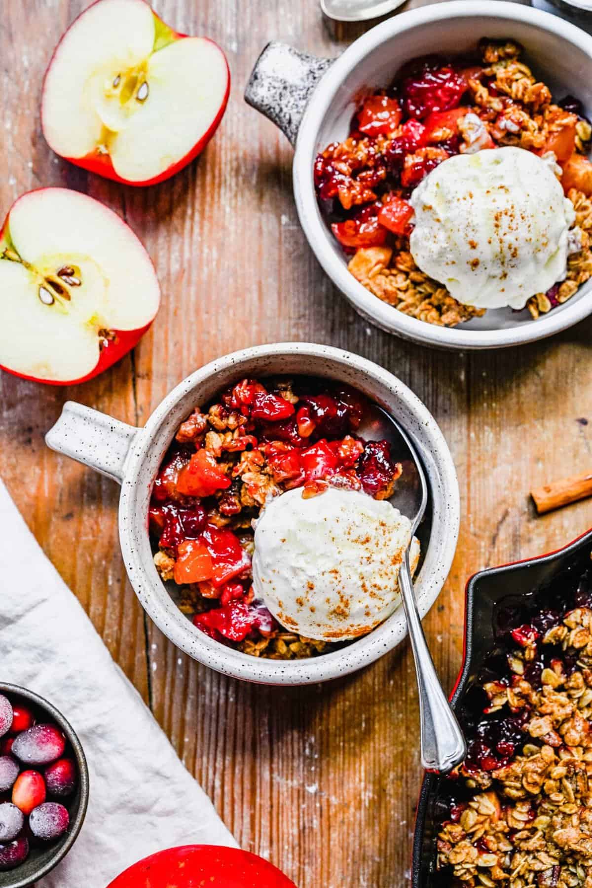 Several servings of apple cranberry crisp in white mugs with ice cream on top sit on a wooden surface. A halved apple sits next to the mugs of apple cranberry crisp.