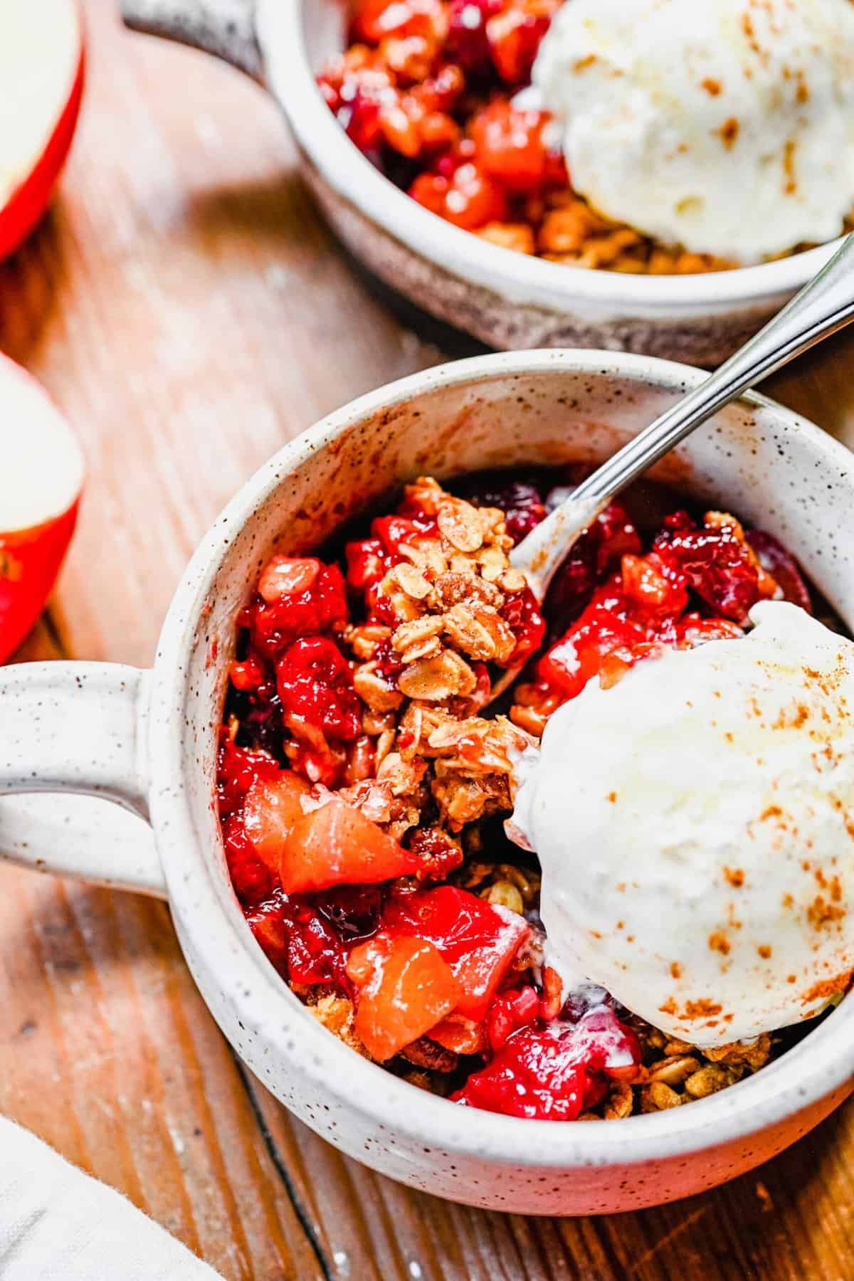 A serving of apple cranberry crisp in a white mug with a scoop of ice cream on top.