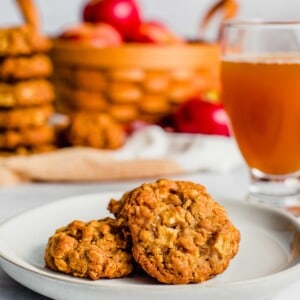 Several golden baked apple crisp cookies sit on a small plate with a basket of apples and a pitcher of apple cider in the background.