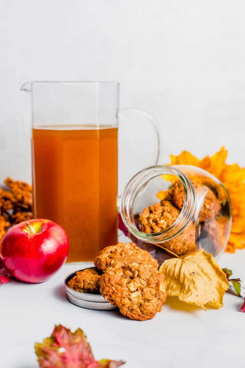 Apple crisp cookies spill from a glass cookie jar and a pitcher of apple cider sits beside them.