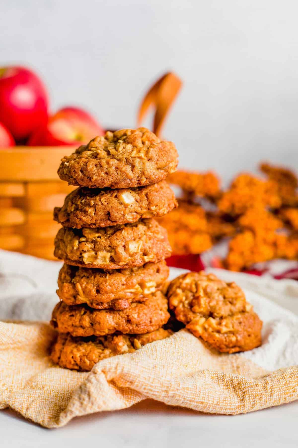 Six apple crisp cookies are stacked with a basket of whole apples in the background.