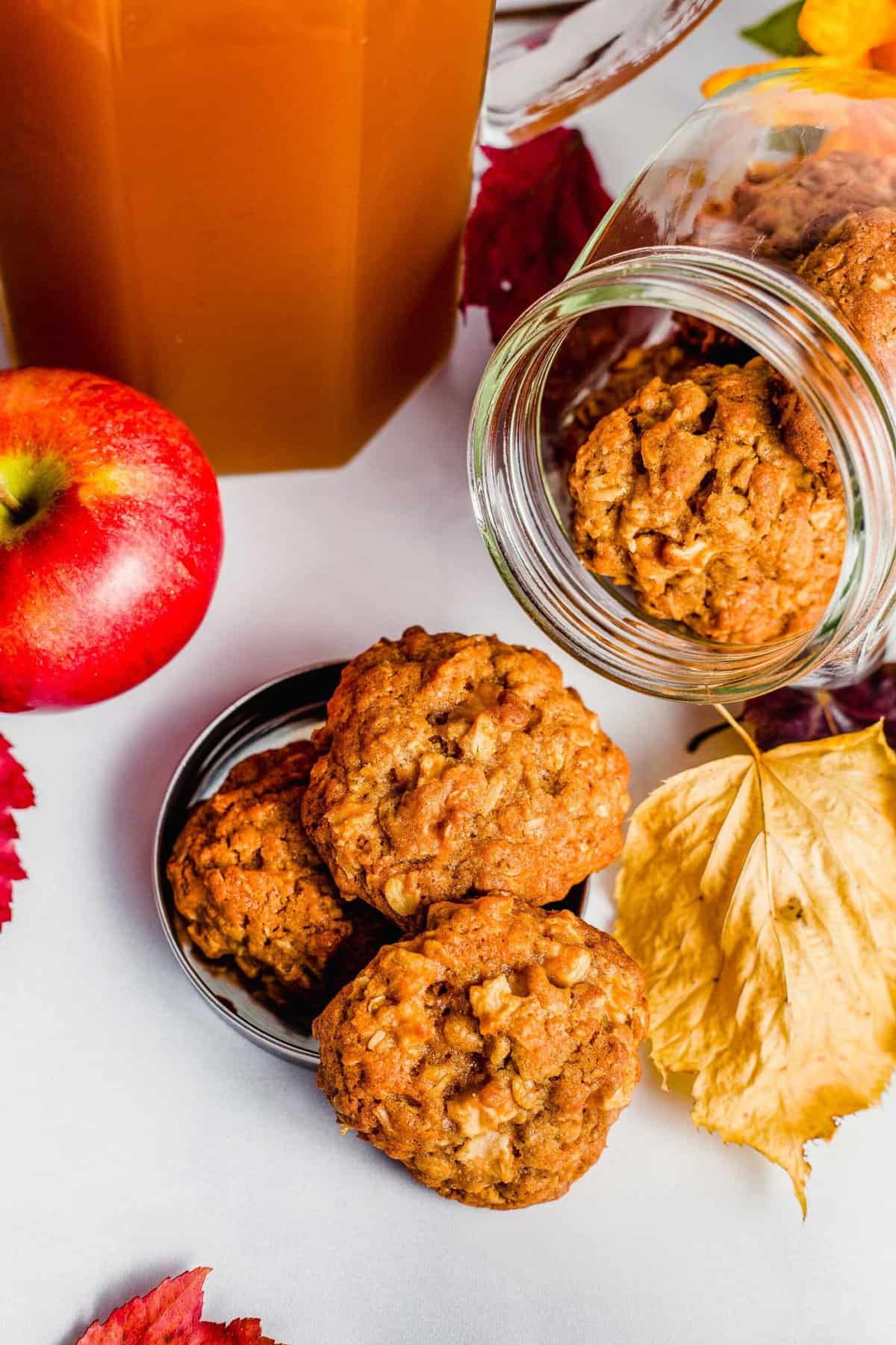 Apple Crisp cookies spill out of a cookie jar amidst fall decor of leaves and whole apples.