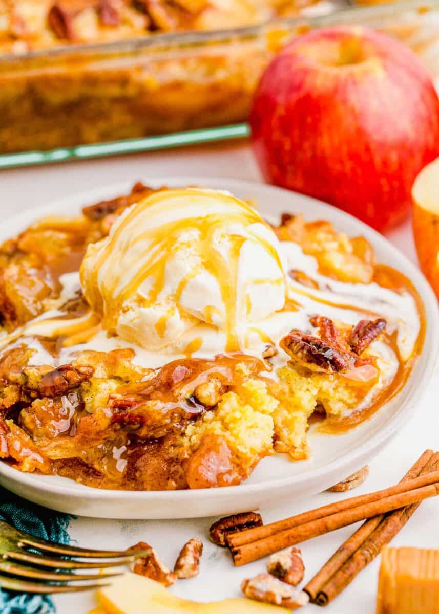 A white plate of Caramel Apple Dump Cake is topped with melting vanilla ice cream and a swirl of caramel sauce drips over the cake. Whole cinnamon sticks, pecans, and a fork lie next to the plate.