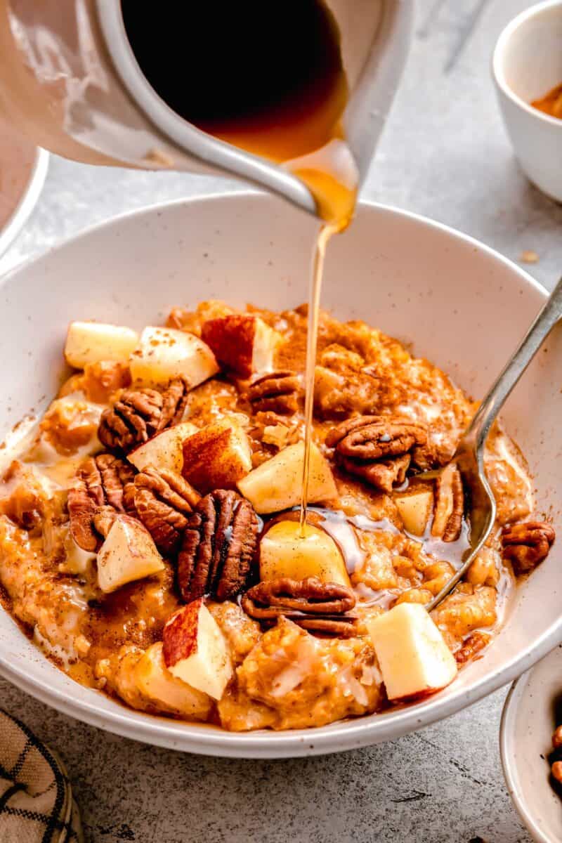 maple syrup being poured on apple cinnamon oats in a white bowl
