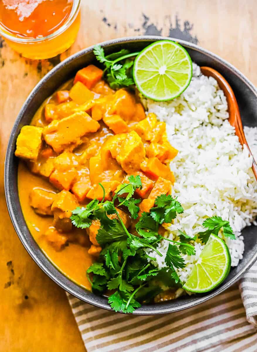 White rice and pumpkin curry are served with cilantro and lime wedges in a black bowl. 
