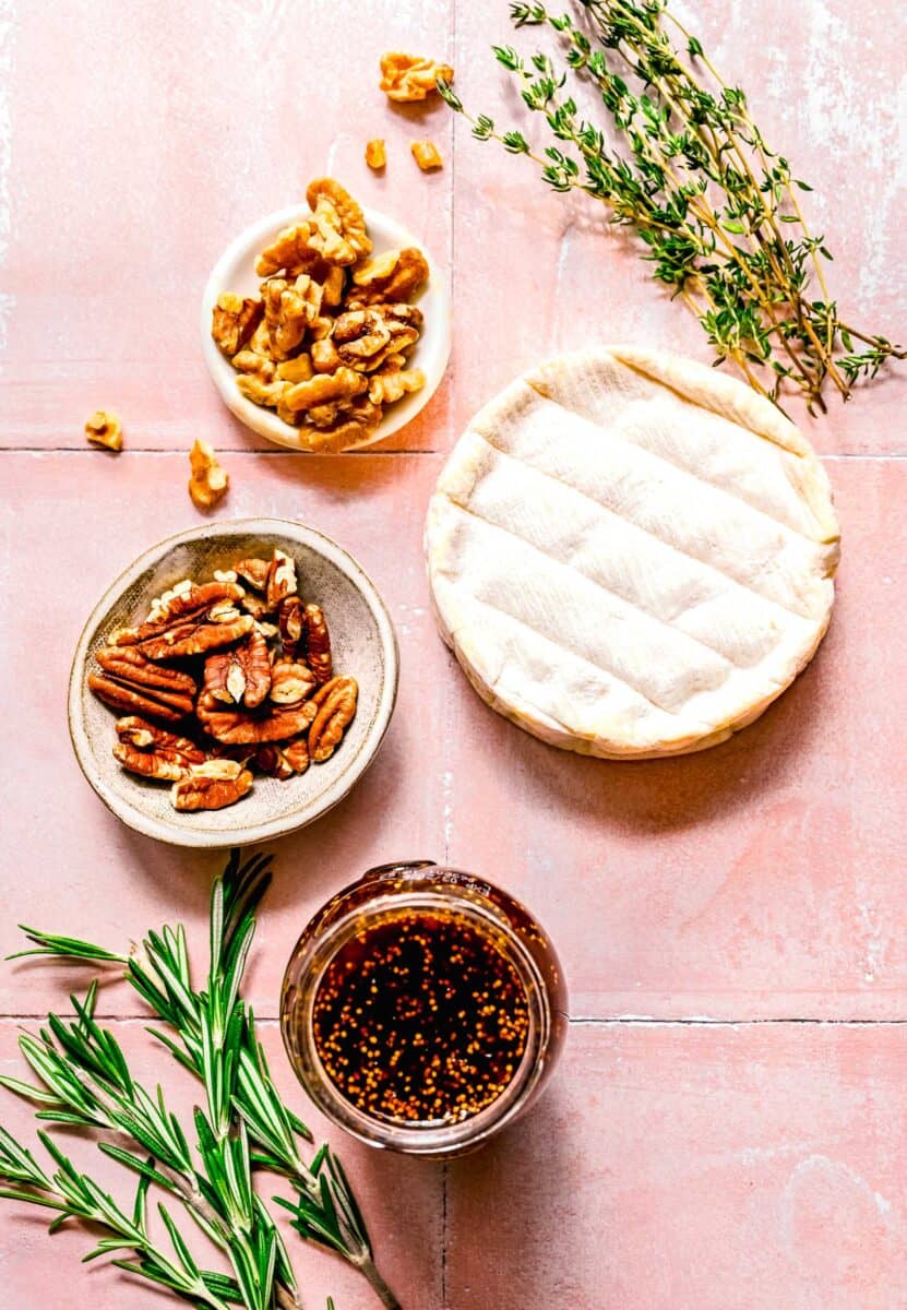 overhead shot of ingredients for baked brie with fig jam, including brie, nuts, fresh thyme, fresh rosemary, and fig jam