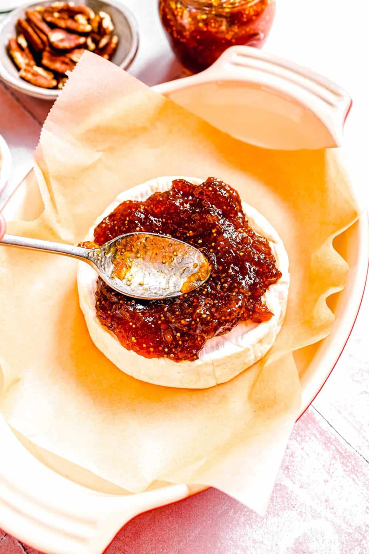 image of fig jam being spread on top of wheel of baked brie in a baking dish