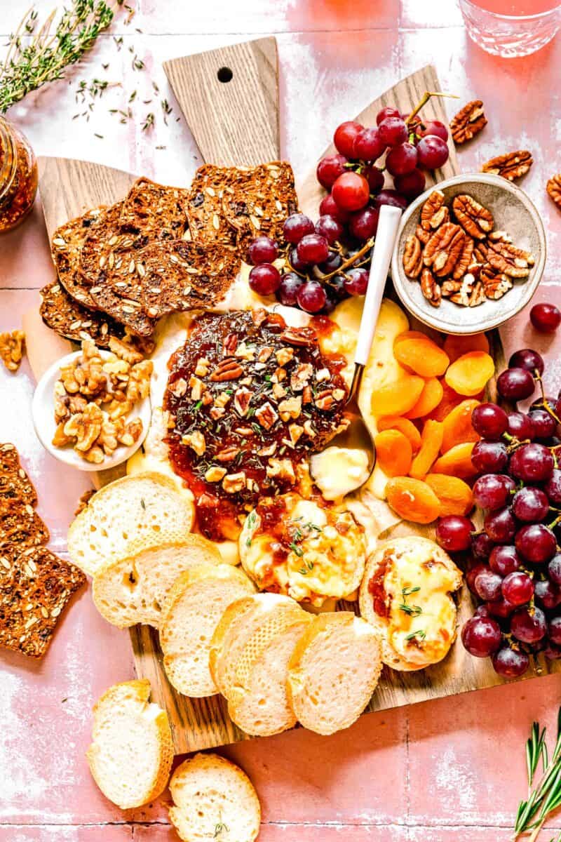 overhead shot of baked brie with fig jam on a cutting board with bread, crackers, nuts, dried fruit, and fresh grapes.