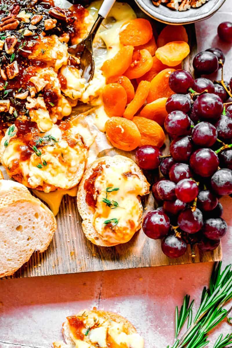 overhead shot of melted baked brie and fig jam spread on top of a slice of small bread. dried fruit and fresh grapes can be seen as well.