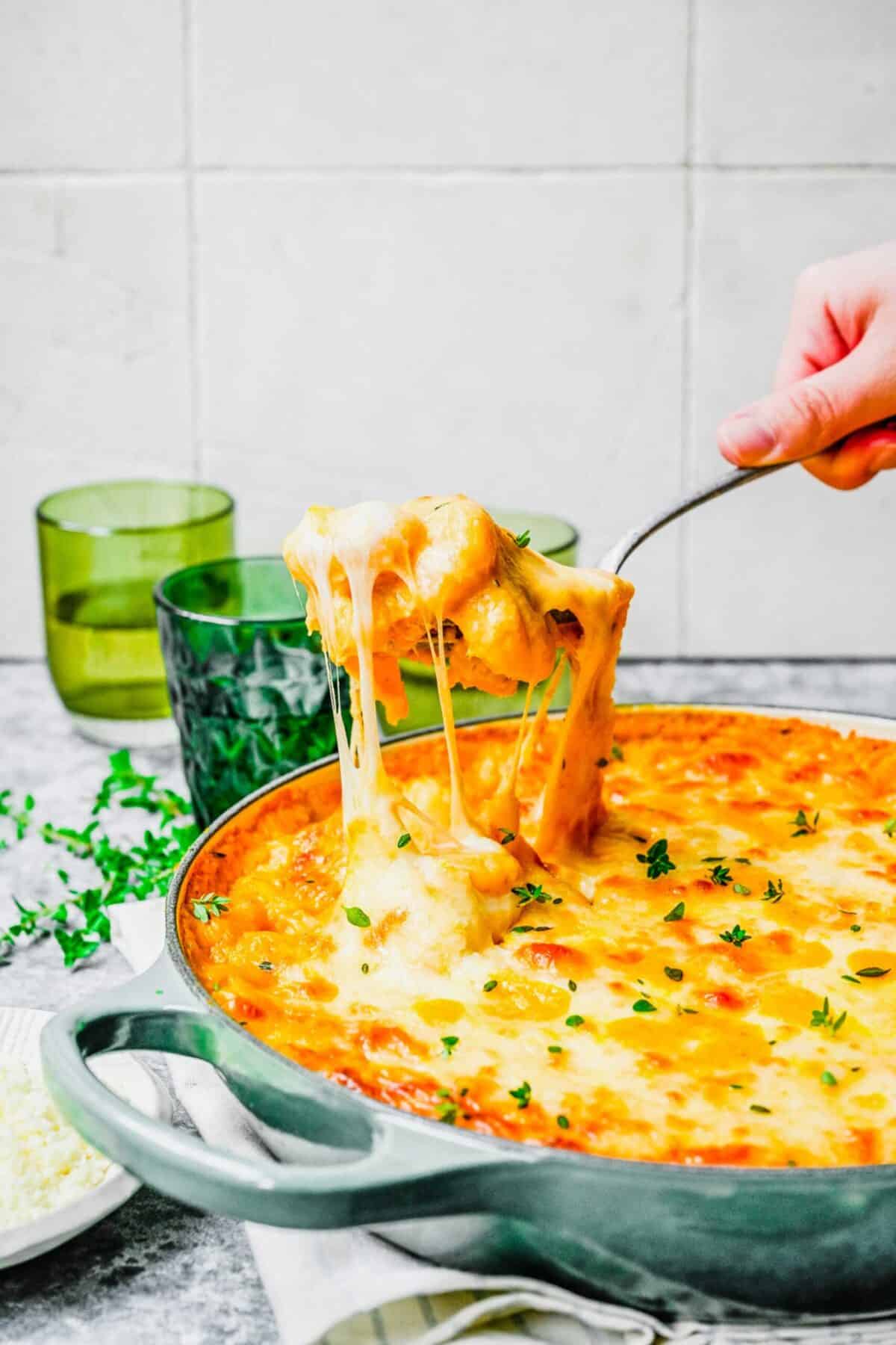 Baked pumpkin gnocchi in a pan, garnished with fresh thyme, with a serving spoon pulling a spoonful out as the cheese stretches, with water glasses in the background