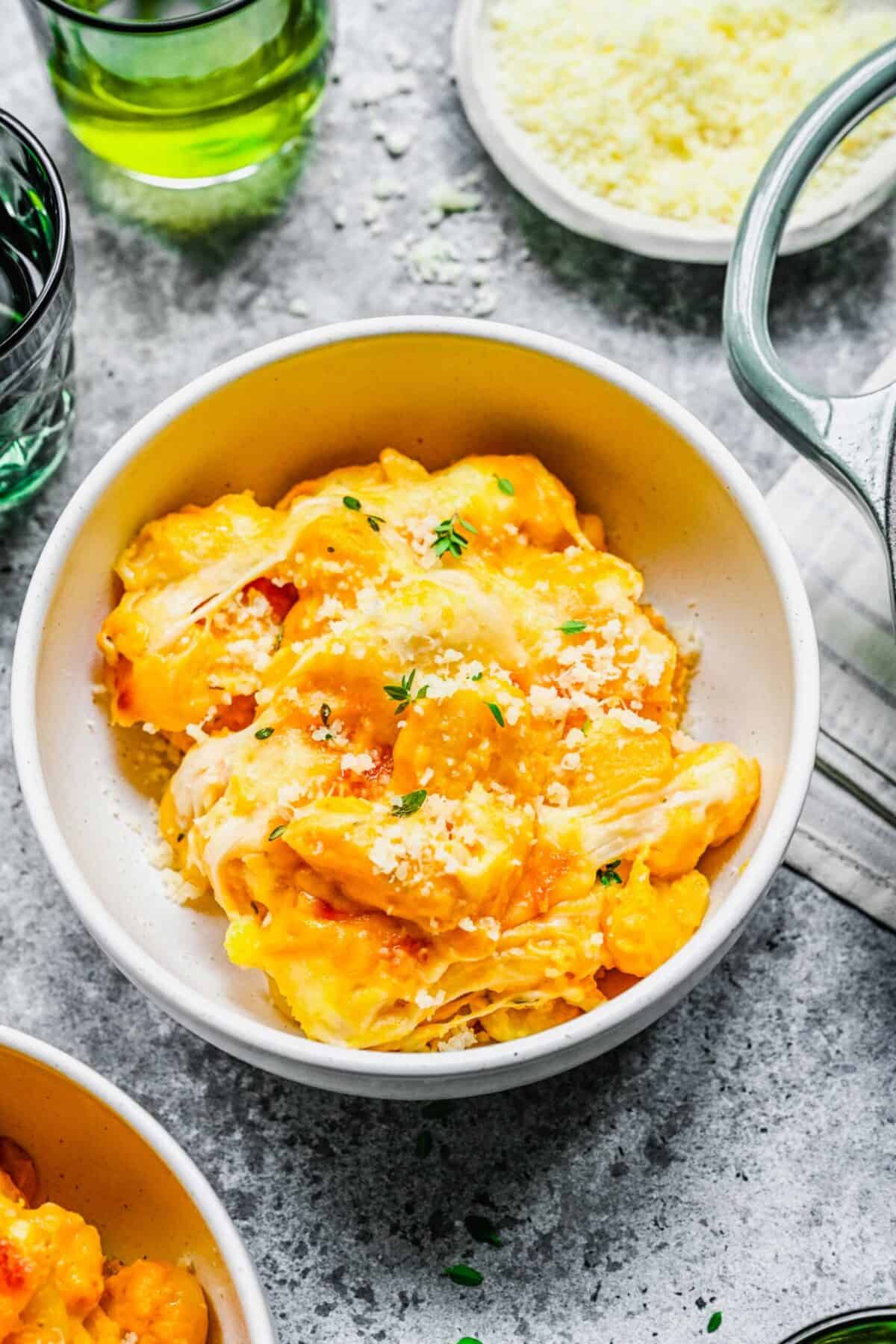 A bowl of baked pumpkin gnocchi topped with fresh thyme, next to another bowl of baked gnocchi and a bowl of shredded parmesan