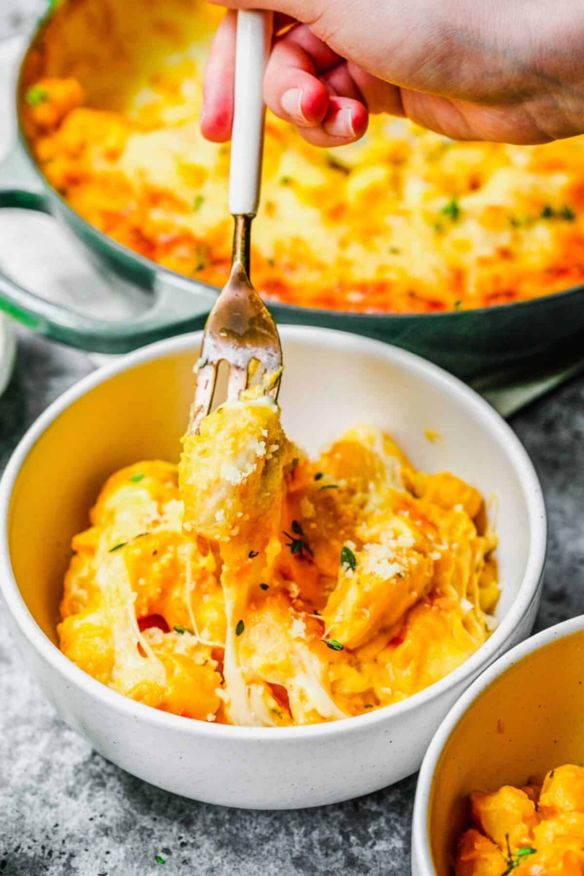 A bowl of baked pumpkin gnocchi with a fork taking a bite out, with a pot of baked gnocchi in the background