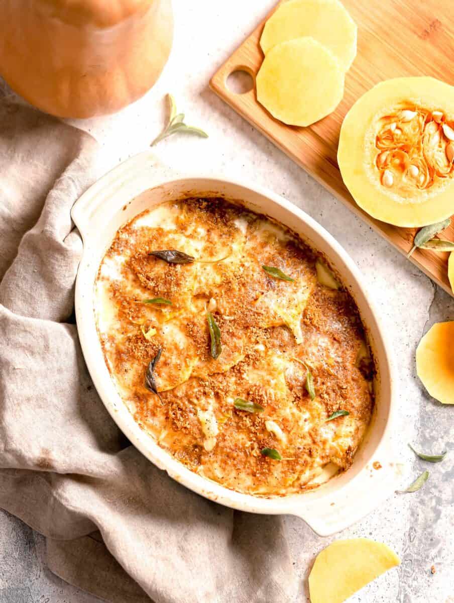 overhead shot of butternut squash gratin out of the oven with uncooked butternut squash on a cutting board.