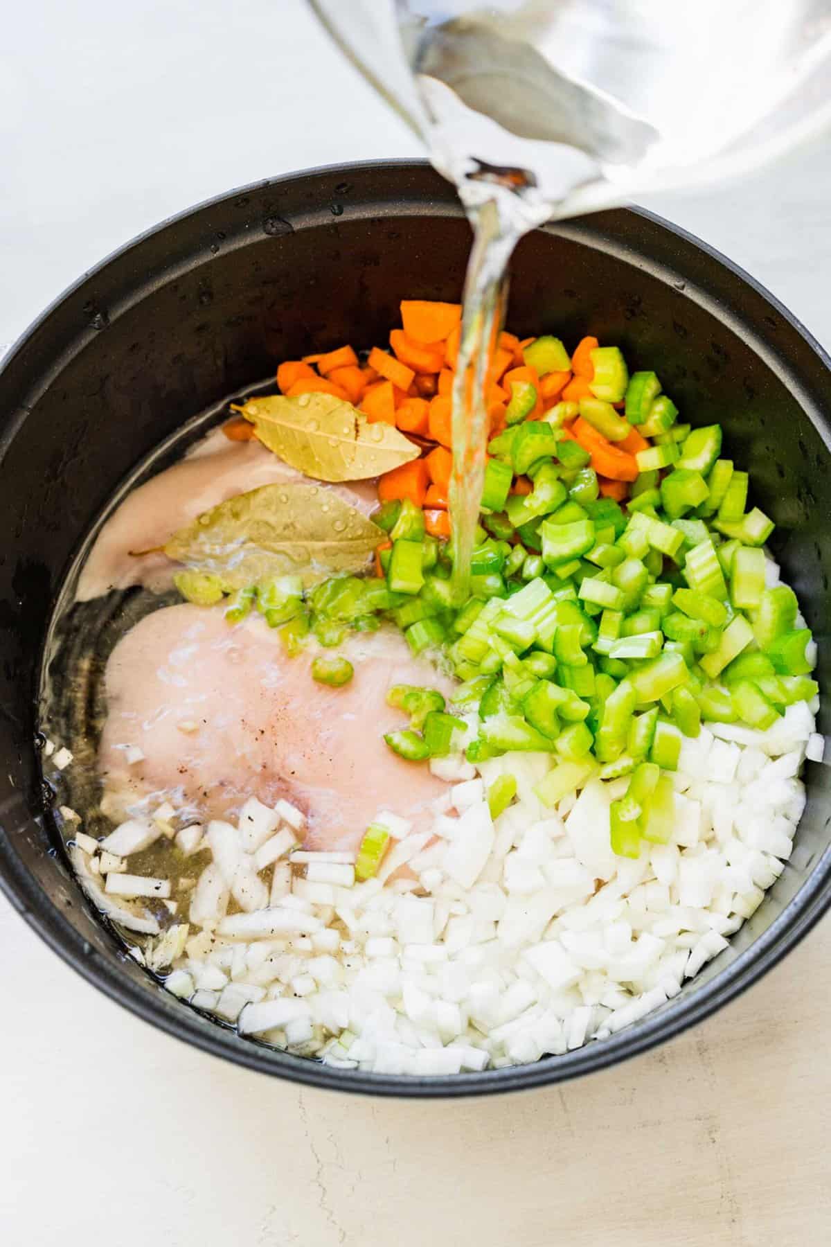 water being poured onto the raw chicken, bay leaves, uncooked carrots, celery, onion, and seasonings in a large pot
