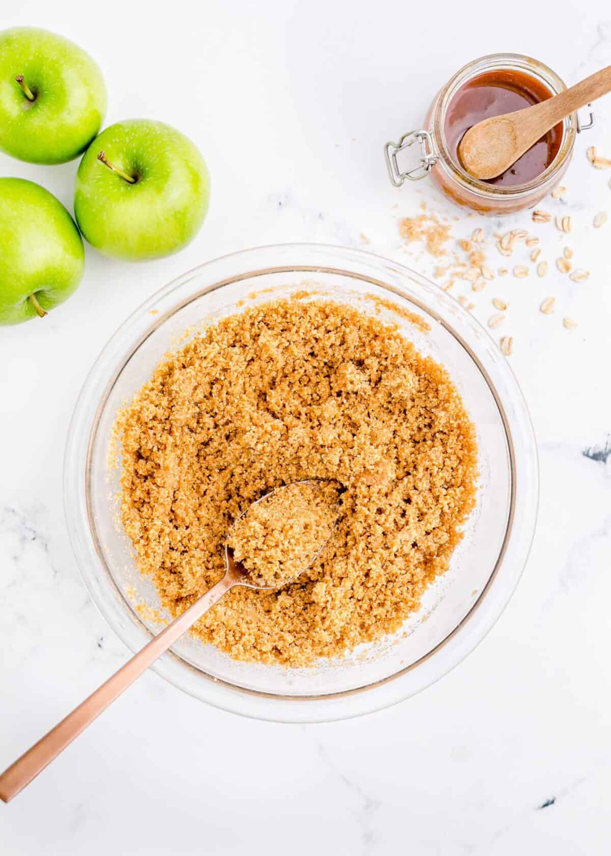A mixing bowl filled with flour, brown sugar, rolled oats, cinnamon, and butter, with a mixing spoon in it, and three apples to the side