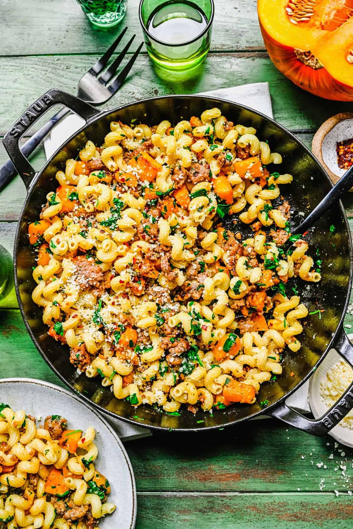 overhead shot of finished pasta with pumpkin and sausage in a staub cast iron pot with a spoon in the pot that has just plated a grey ceramic dish with some pasta.