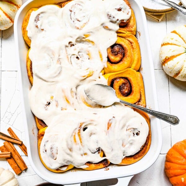 A baking dish of cinnamon rolls, mostly covered with frosting, with a frosting covered spoon on top, next to some pumpkins and cinnamon sticks