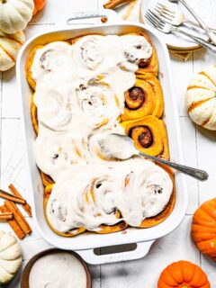 A baking dish of cinnamon rolls, mostly covered with frosting, with a frosting covered spoon on top, next to some pumpkins and cinnamon sticks