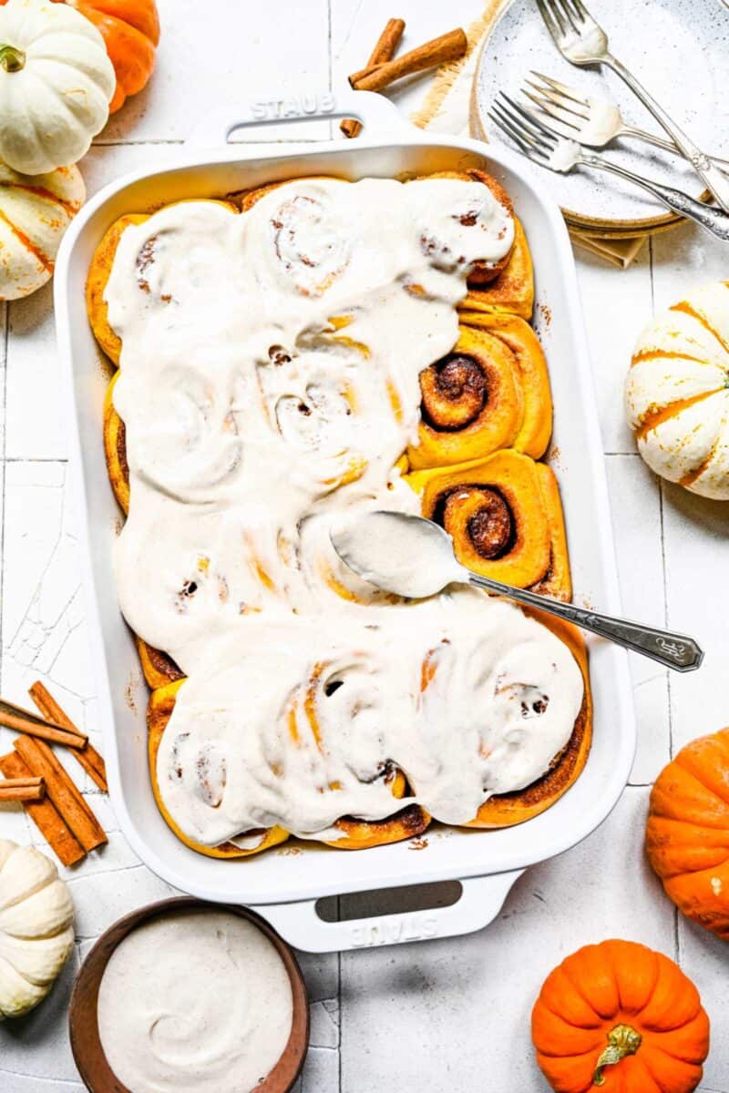 A baking dish of cinnamon rolls, mostly covered with frosting, with a frosting covered spoon on top, next to some pumpkins and cinnamon sticks