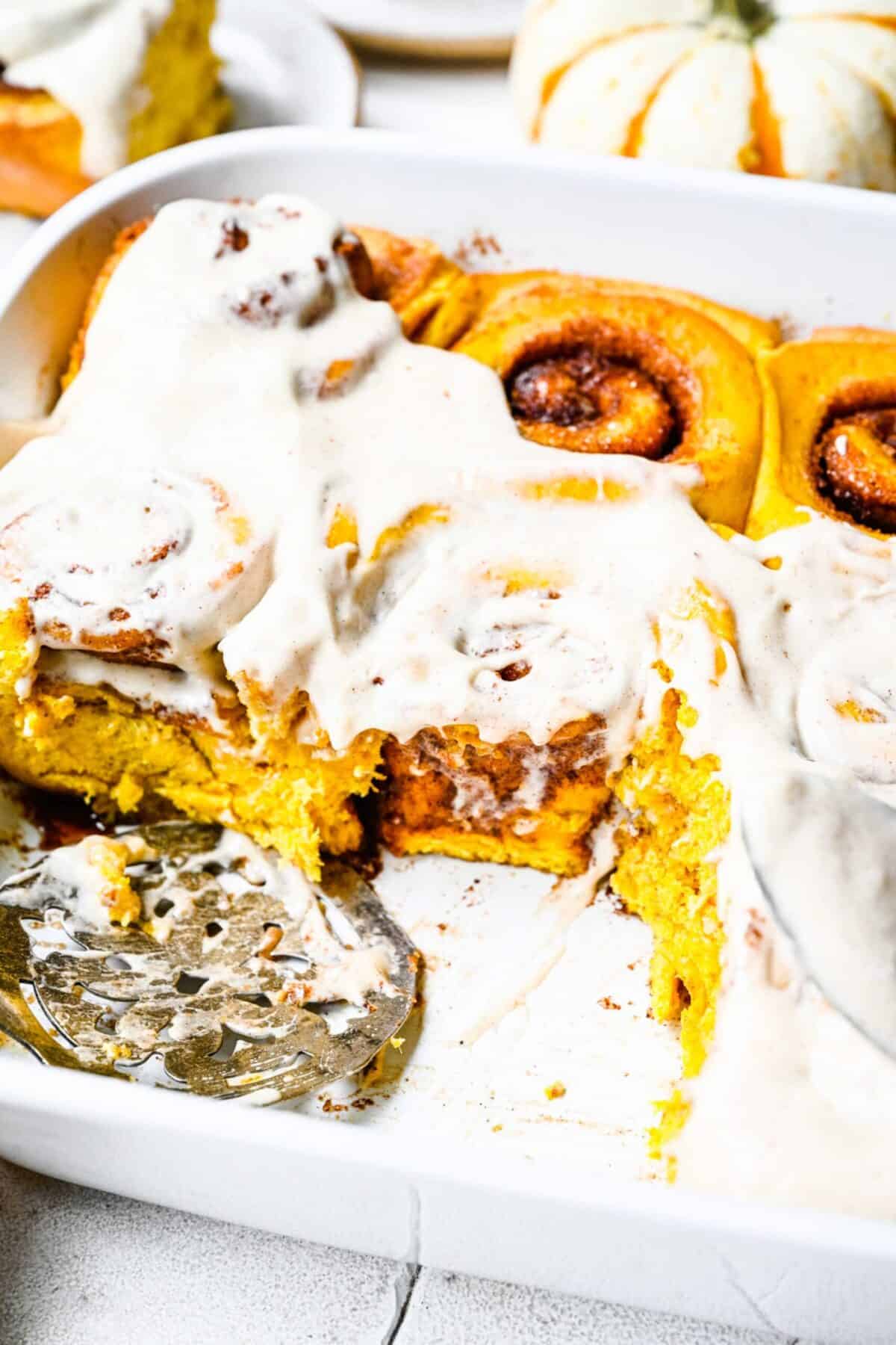 Close up of a some cinnamon rolls in a baking tray, covered in frosting, with a serving spoon