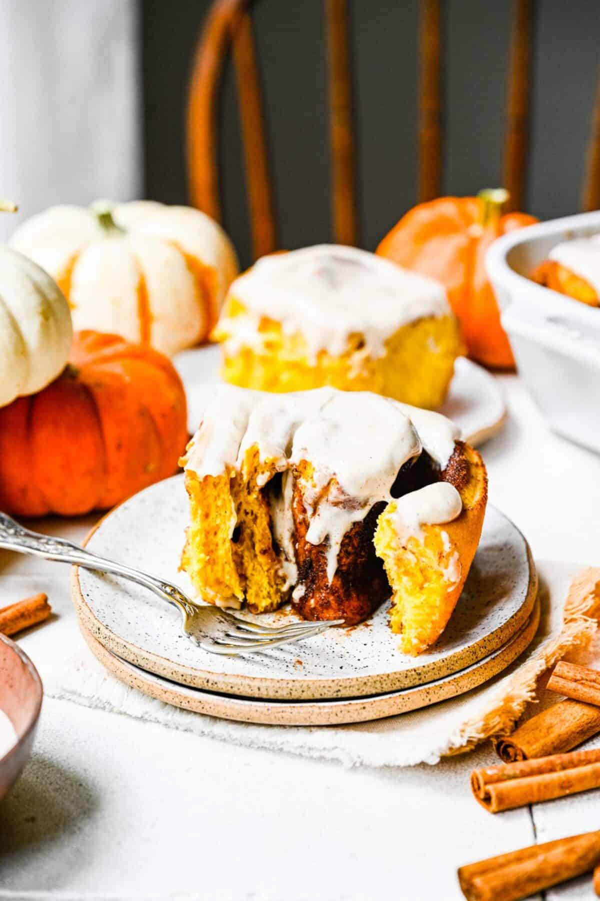 A pumpkin cinnamon roll on a plate, with frosting on top, and a fork on the plate, with pumpkins in the background