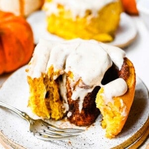 Close up of a pumpkin cinnamon roll on a plate with a fork, with another cinnamon roll on a plate in the background